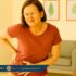 Middle-aged woman pressing her hip and abdomen in visible discomfort, standing in a bright, wellness-themed room with indoor plants and soft decor.