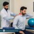 Pakistani physiotherapist in white coat examining a seated patient with neck and shoulder pain in a modern clinic.
