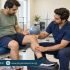 Male physiotherapist examining a seated patient’s bent knee while the foot rests on a padded stool.